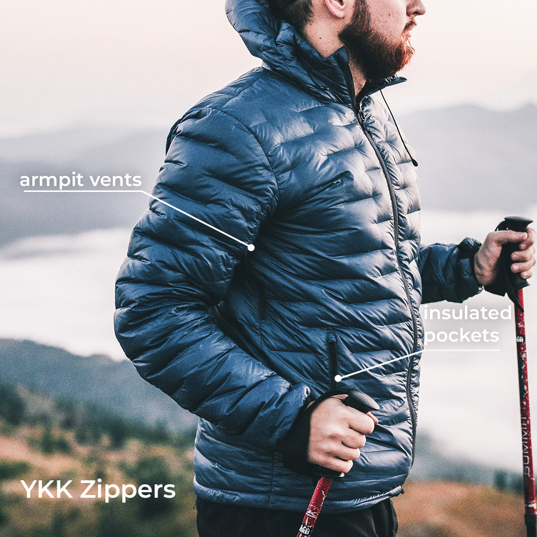 A man wearing a dark blue quilted puffer jacket outdoors on a mountain, with text callouts pointing to armpit vents, insulated pockets, and YKK zippers.