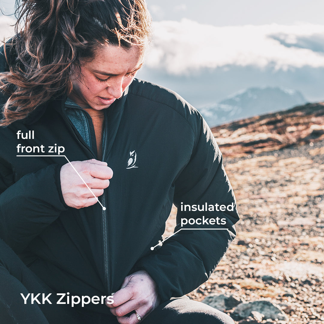 A woman outdoors on a rocky mountain setting wearing a black YKK jacket, demonstrating the full front zip and pointing to insulated pockets with text overlays.