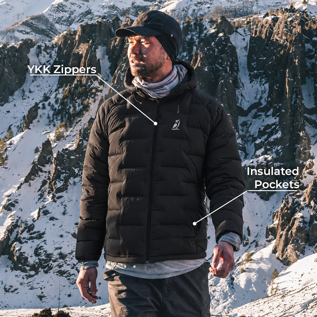 A man wearing an insulated puffer jacket stands in a snowy mountain landscape. White text labels point to the jacket, reading 'YKK Zippers' near the chest zipper area and 'Insulated Pockets' near the lower pocket area.