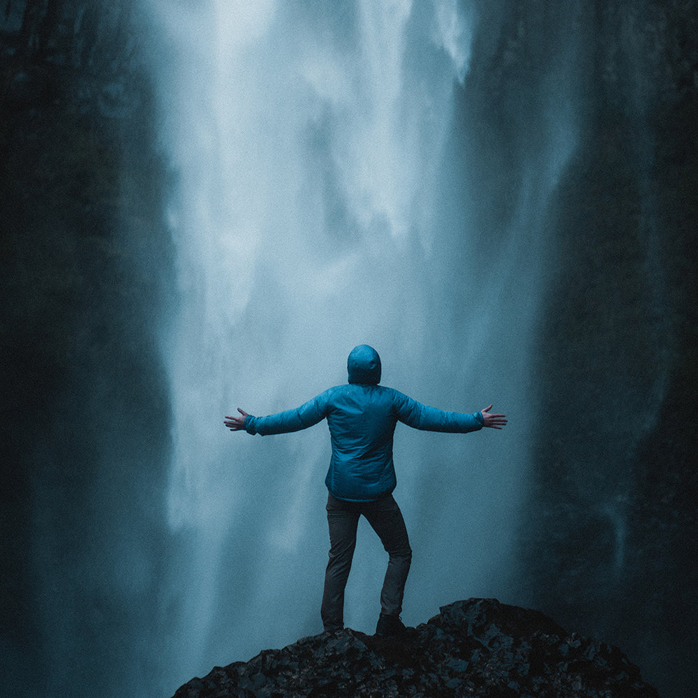 rear view of man wearing blue mid layer hoodie in front of waterfall