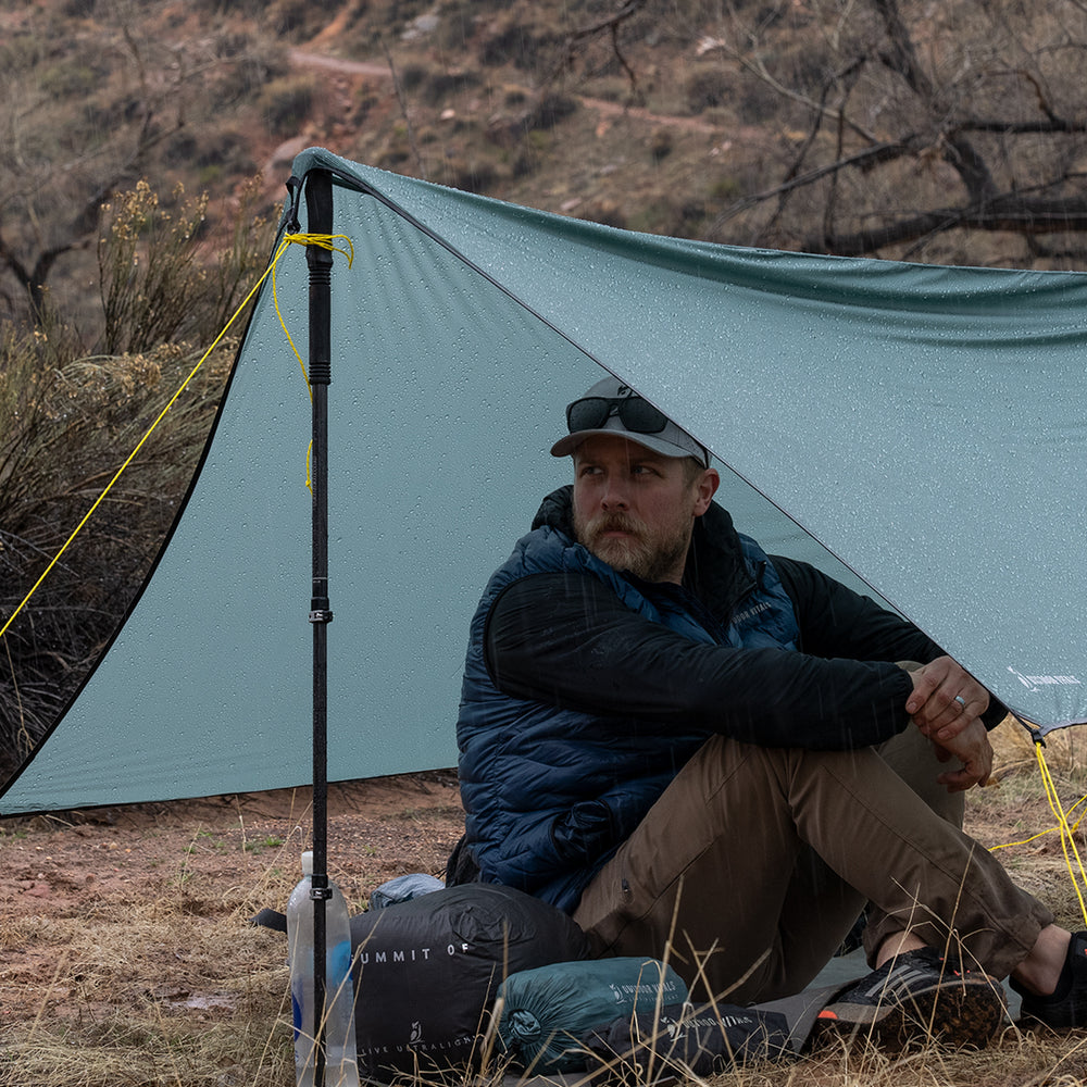 side view of man sitting beneath tarp shelter during rain storm