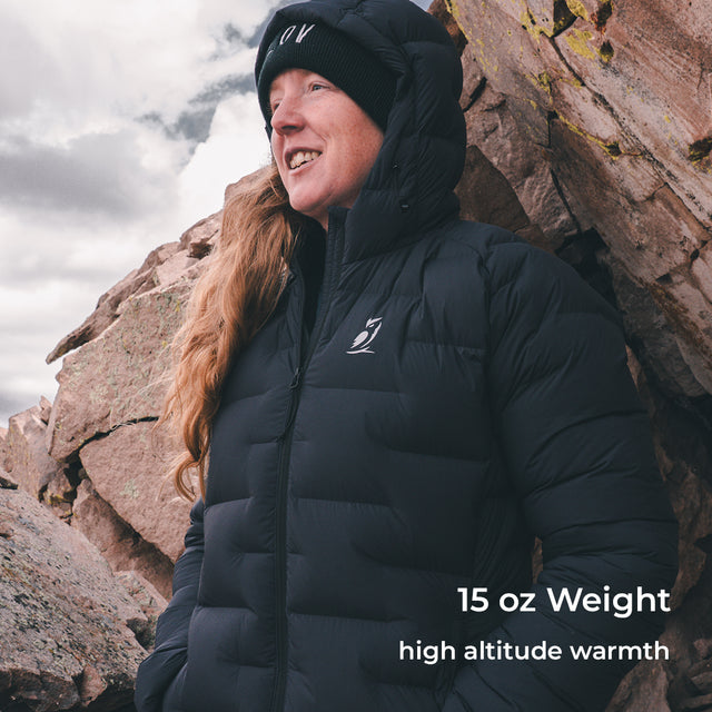 A woman wearing a black hooded puffer jacket and black knit beanie stands outdoors against large rock formations under a cloudy sky. White text in the lower right reads '15 oz Weight high altitude warmth.