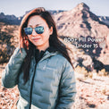 A woman wearing a light blue-gray puffer jacket and reflective sunglasses stands outdoors in a desert mountain landscape. White text on the right reads '800+ Fill Power, Under 15 oz.