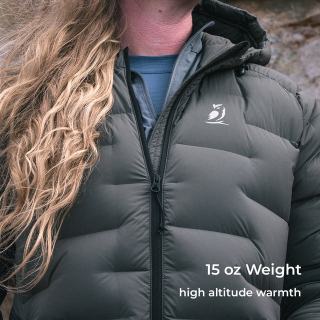 A close-up of a person wearing a dark gray puffer jacket outdoors. The jacket has horizontal quilting, a front zipper, a small white bird logo on the left chest, and text overlay reading '15 oz Weight high altitude warmth'.