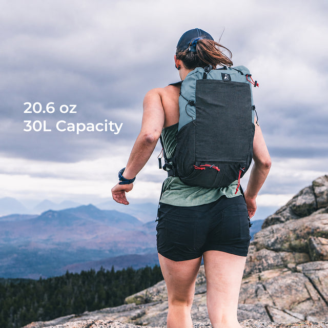 trail runner on mountain peak while wearing blue-gray fastpack. Text callout says "20.6 oz, 30L capacity."