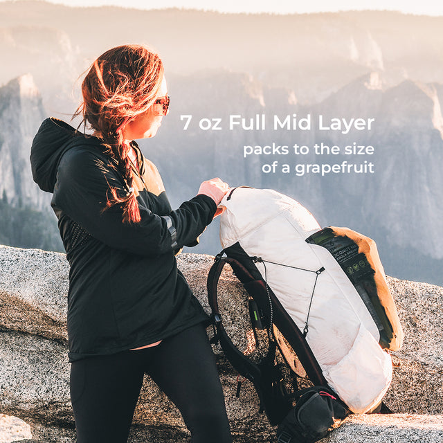 A woman in a black hooded jacket packs a white and black backpack on a rocky mountain ridge. Text on the image reads '7 oz Full Mid Layer packs to the size of a grapefruit'.