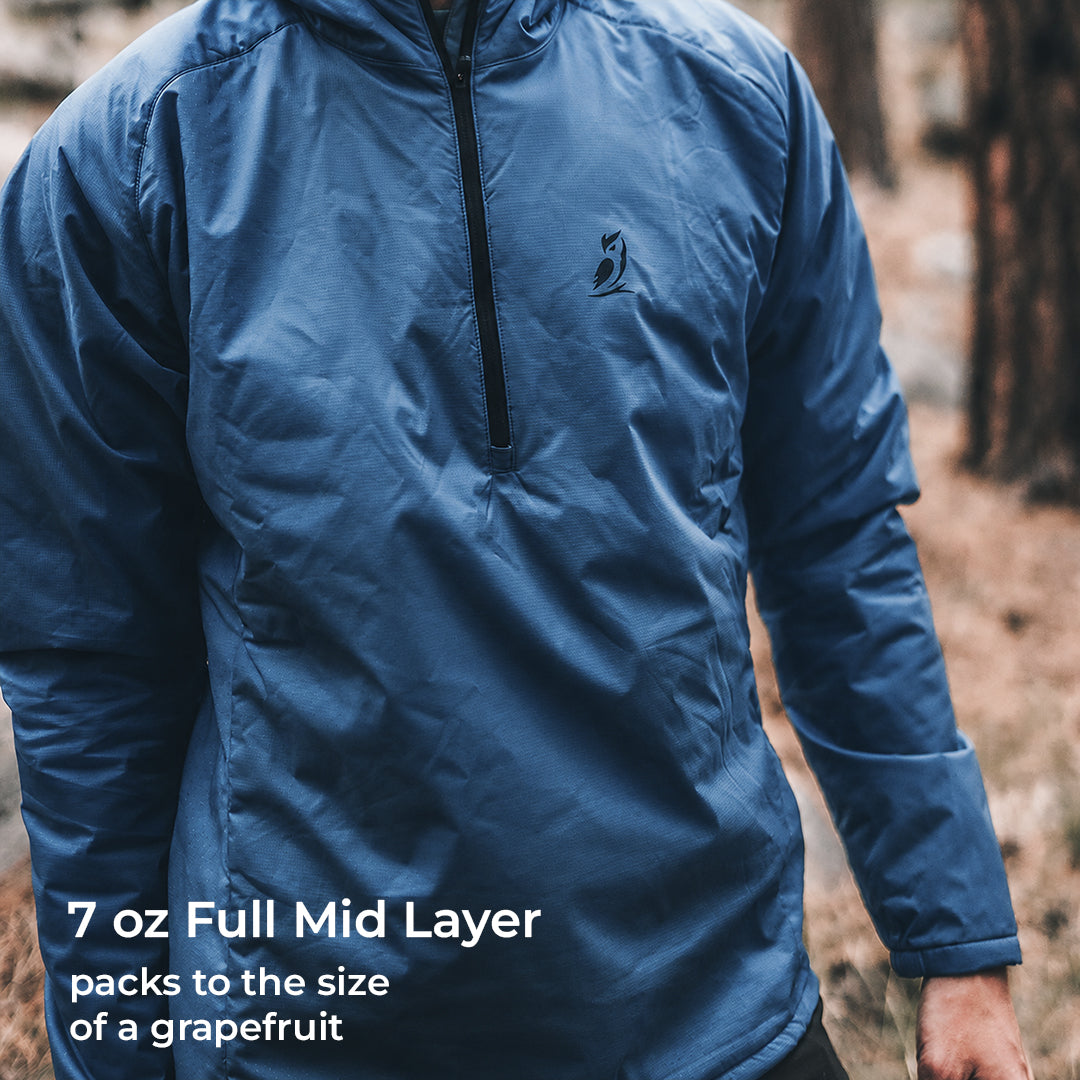 A man wearing a blue lightweight half-zip performance jacket in an outdoor forest setting, with text overlay in the lower left reading '7 oz Full Mid Layer packs to the size of a grapefruit'.