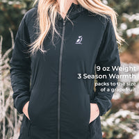 A woman wearing a black lightweight zip-front jacket outdoors, with her hands in the pockets. The jacket has a small white bird logo on the left chest and white text on the right side that reads: '9 oz Weight 3 Season Warmth packs to the size of a grapefruit.