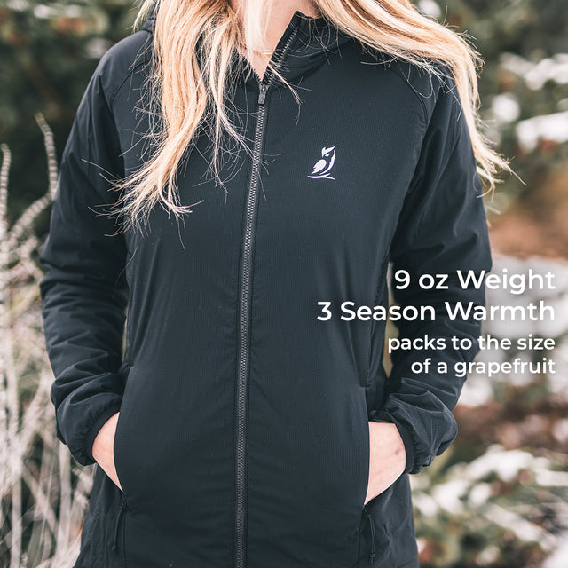 A woman wearing a black lightweight zip-front jacket outdoors, with her hands in the pockets. The jacket has a small white bird logo on the left chest and white text on the right side that reads: '9 oz Weight 3 Season Warmth packs to the size of a grapefruit.