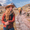 A woman hiking in a mountainous outdoor setting, wearing a wide-brim straw hat, rust-red jacket, and black backpack. White text in the upper right reads: '9 oz Weight 3 Season Warmth packs to the size of a grapefruit'.