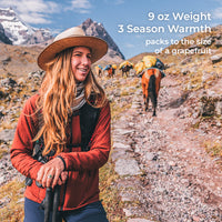 A woman hiking in a mountainous outdoor setting, wearing a wide-brim straw hat, rust-red jacket, and black backpack. White text in the upper right reads: '9 oz Weight 3 Season Warmth packs to the size of a grapefruit'.