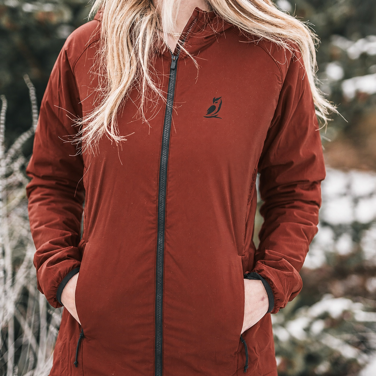 A woman wearing a rust-brown hooded zip-up jacket with a small black bird logo on the left chest, standing outdoors in a snowy, blurred environment.