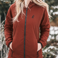 A woman wearing a rust-brown hooded zip-up jacket with a small black bird logo on the left chest, standing outdoors in a snowy, blurred environment.