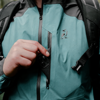 Close-up of a person wearing a waterproof jacket with a black backpack, opening a zippered pocket on the chest.