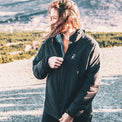 A woman wearing a black and gray color-blocked athletic jacket with a small white logo on the left chest, standing outdoors in a gravel area with mountains in the background.