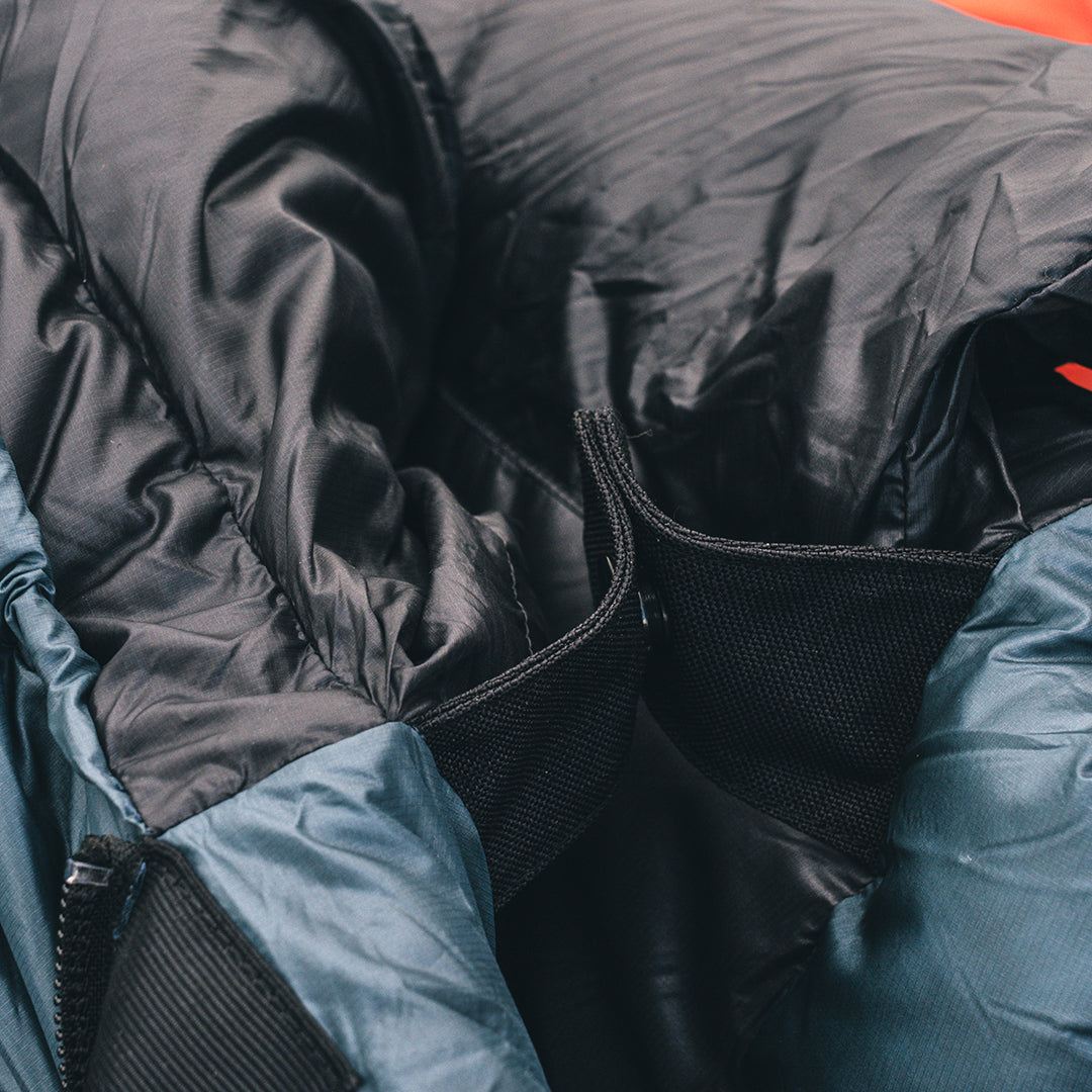 Close-up detail of a backpacking sleeping bag showing the black insulated fill, black zipper track, black snap closure at the collar, and blue-gray shell fabric.