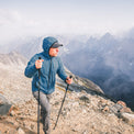 A man wearing a blue hooded jacket, gray pants, and a black cap hikes with trekking poles on a rocky mountain ridge with a dramatic mountain range in the background.