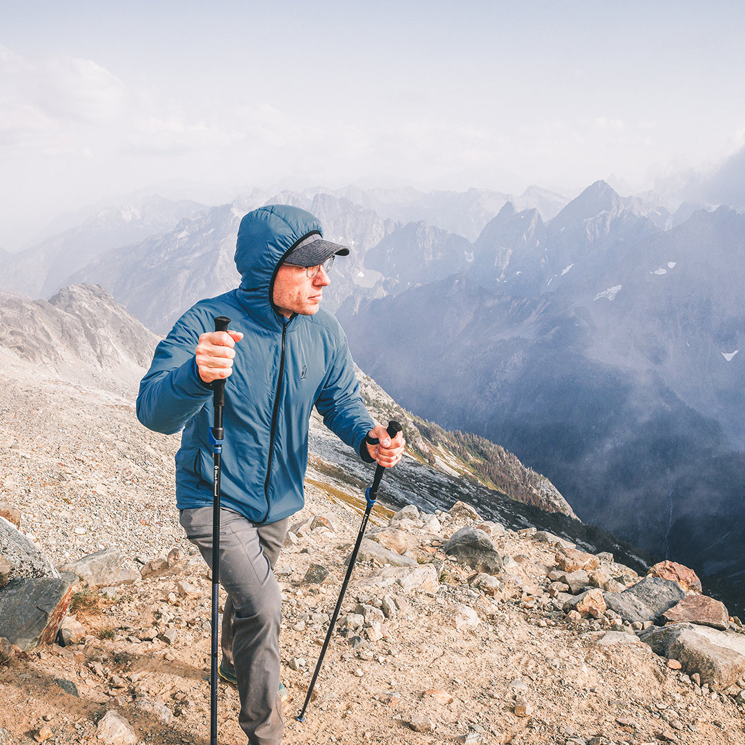 A man wearing a blue hooded jacket, gray pants, and a black cap hikes with trekking poles on a rocky mountain ridge with a dramatic mountain range in the background.