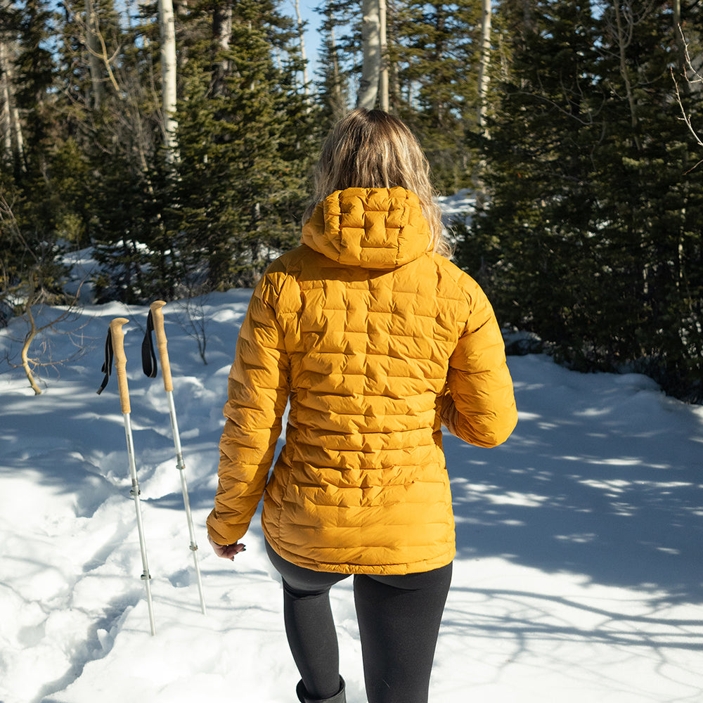 Woman wearing insulated hooded down jacket from the back while walking through snowy forest terrain.