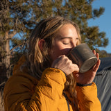 Woman wearing insulated down jacket drinking from a camp mug outdoors in cold conditions.