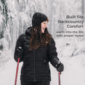 A woman wearing a black puffer jacket, black beanie, and black gloves is skiing in the snow, holding ski poles. Text on the right reads 'Built For Backcountry Comfort' and 'warm into the 30s with proper layers'.
