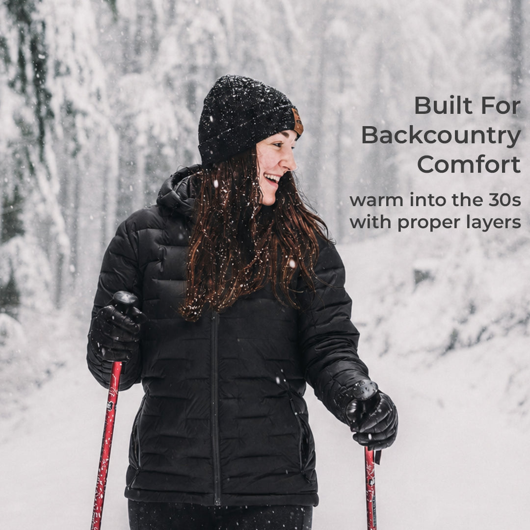 A woman wearing a black puffer jacket, black beanie, and black gloves is skiing in the snow, holding ski poles. Text on the right reads 'Built For Backcountry Comfort' and 'warm into the 30s with proper layers'.