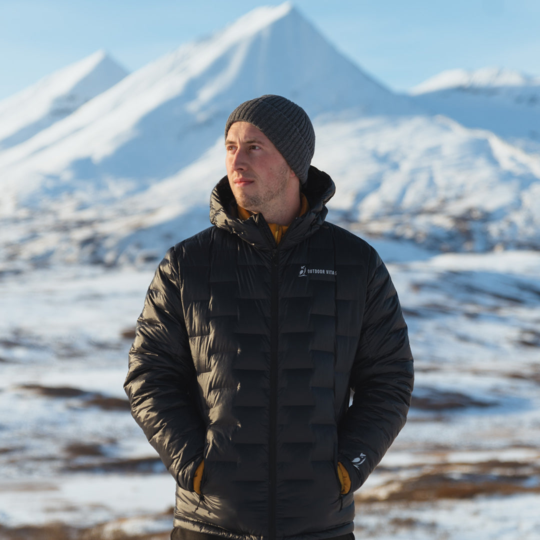 Person wearing a black Outdoor Vitals insulated jacket and gray knit beanie, standing with hands in pockets in front of a snow-covered mountain range under clear blue skies.