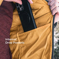 Close-up of a person's hands opening a mustard yellow puffer jacket to reveal an internal drop pocket holding a black smartphone. White text on the image reads 'Internal Drop Pockets'.