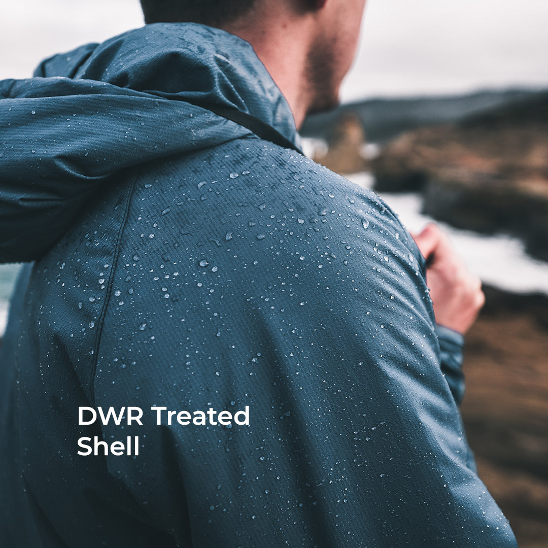 A man viewed from behind wearing a dark blue-gray hooded jacket covered in water droplets, standing outdoors near the water with the text 'DWR Treated Shell' overlaid on the lower left.
