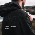 A man wearing a black hooded jacket outdoors, shown from the back with water droplets on the treated shell. The text 'DWR Treated Shell' is overlaid on the lower left, with a blurred coastal background.