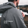 A man viewed from behind wearing a dark gray hooded jacket covered in water droplets, standing outdoors near the ocean with the text 'DWR Treated Shell' overlaid on the lower left.