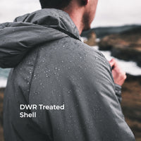A man viewed from behind wearing a dark gray hooded jacket covered in water droplets, standing outdoors near the ocean with the text 'DWR Treated Shell' overlaid on the lower left.