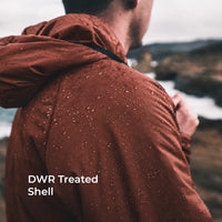 A man seen from behind wearing a hooded jacket covered in water droplets, standing outdoors near a rocky coastal landscape. White text on the lower left reads 'DWR Treated Shell'.