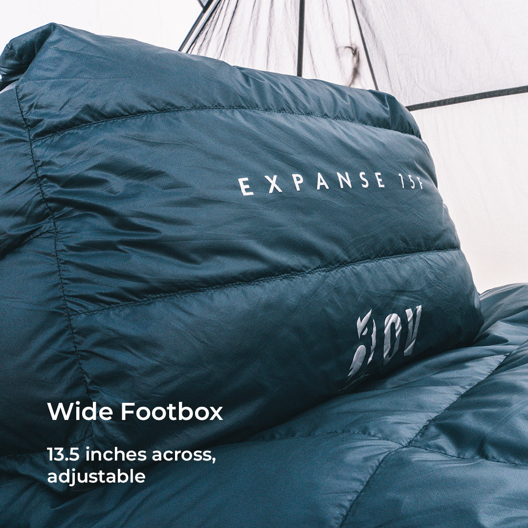 A close-up of a dark blue down sleeping bag inside a tent, showing white printed branding and text that reads 'Wide Footbox 13.5 inches across, adjustable'.