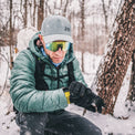 A man in winter gear, including a puffer jacket, black gloves, a gray cap, and mirrored ski goggles, checks his smartwatch while kneeling in the snow next to a tree trunk.