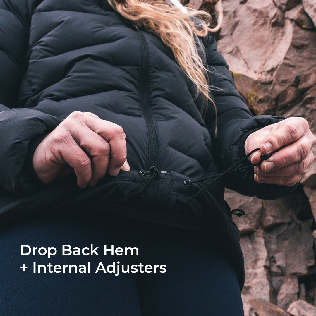 A close-up of a person wearing a black puffer jacket outdoors, adjusting the hem with internal hardware. White text in the lower left reads 'Drop Back Hem + Internal Adjusters'.