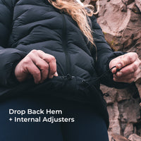 A close-up of a person wearing a black puffer jacket outdoors, adjusting the hem with internal hardware. White text in the lower left reads 'Drop Back Hem + Internal Adjusters'.