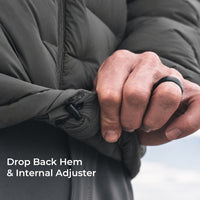 Close-up of a person's hands adjusting the hem of a dark gray puffer jacket, demonstrating the drop back hem and internal adjuster feature. A black ring is worn on one finger, and white text in the lower-left corner reads 'Drop Back Hem & Internal Adjuster'.