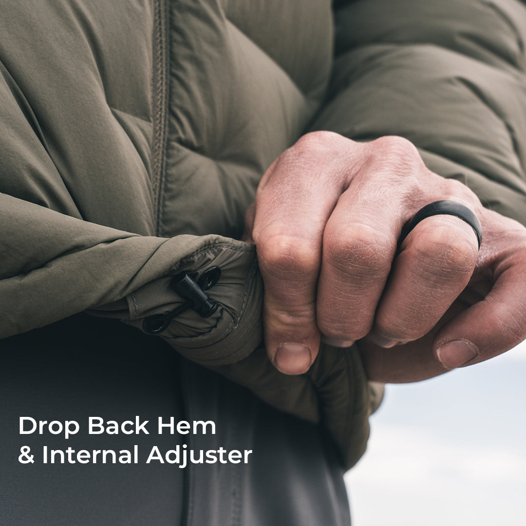 Close-up of a person's hands adjusting the hem of a dark green puffer jacket, demonstrating the drop back hem and internal adjuster feature. A black ring is worn on one finger, and white text in the lower-left corner reads 'Drop Back Hem & Internal Adjuster'.