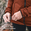 Close-up of a person wearing an rust-brown lightweight jacket and adjusting the back hem with an internal elastic adjuster. White text in the lower left reads 'Drop Back Hem & Internal Adjuster'.