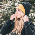 A woman wearing a fitted black hooded jacket and a mustard yellow Carhartt beanie, with the Carhartt logo visible on the brim. She is outdoors in front of blurred evergreen trees, adjusting the hood of the jacket.