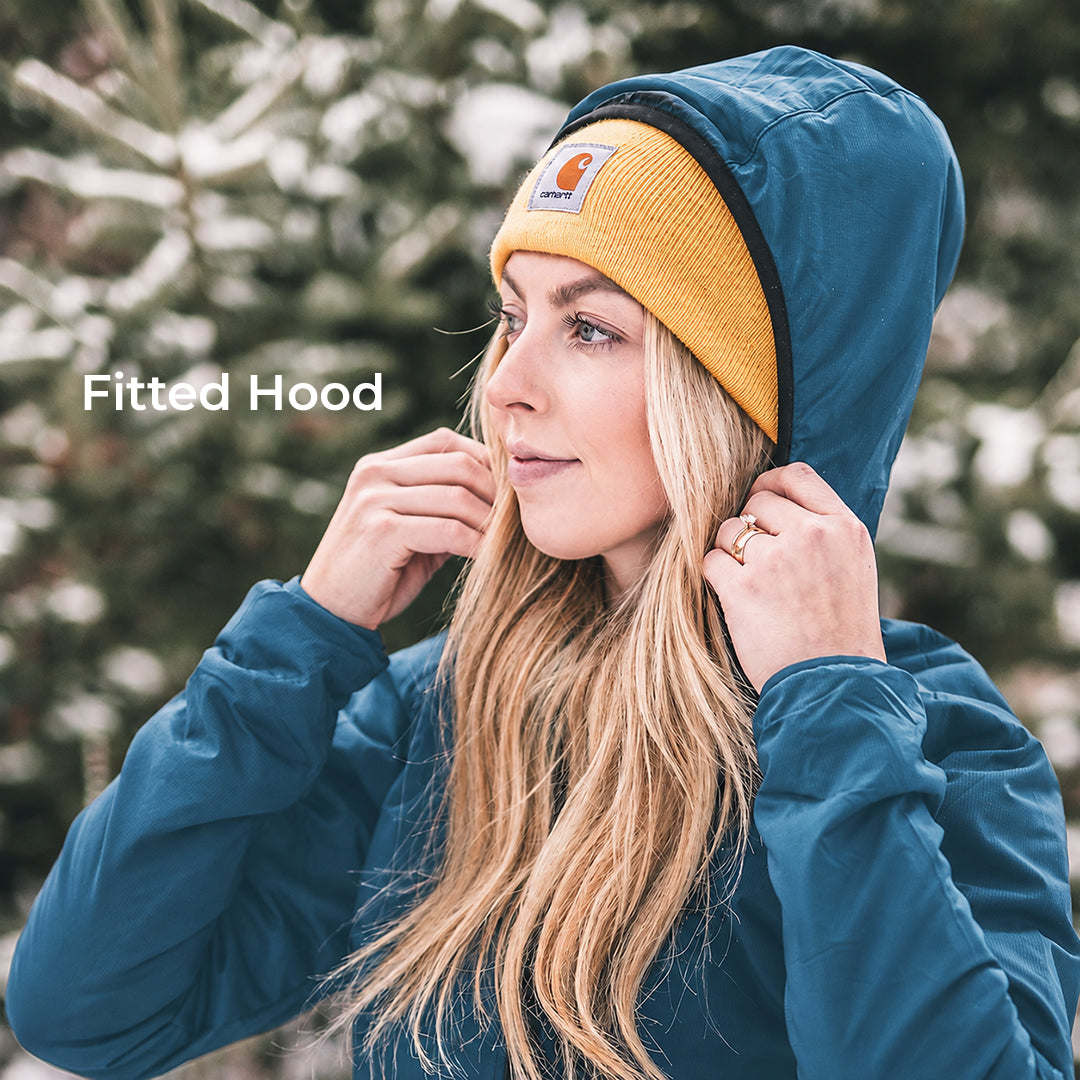 A woman wearing a fitted teal blue hooded jacket with a mustard yellow Carhartt knit beanie, posing outdoors in front of snowy pine trees. The text 'Fitted Hood' is overlaid on the left side.