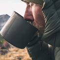 A close-up lifestyle shot of a man wearing a puffy jacket and drinking from a metal camping mug outdoors in a mountainous setting.