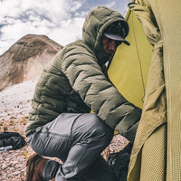 A man in a green puffer jacket and gray pants crouches beside a tent in a rocky mountain setting under a cloudy sky.