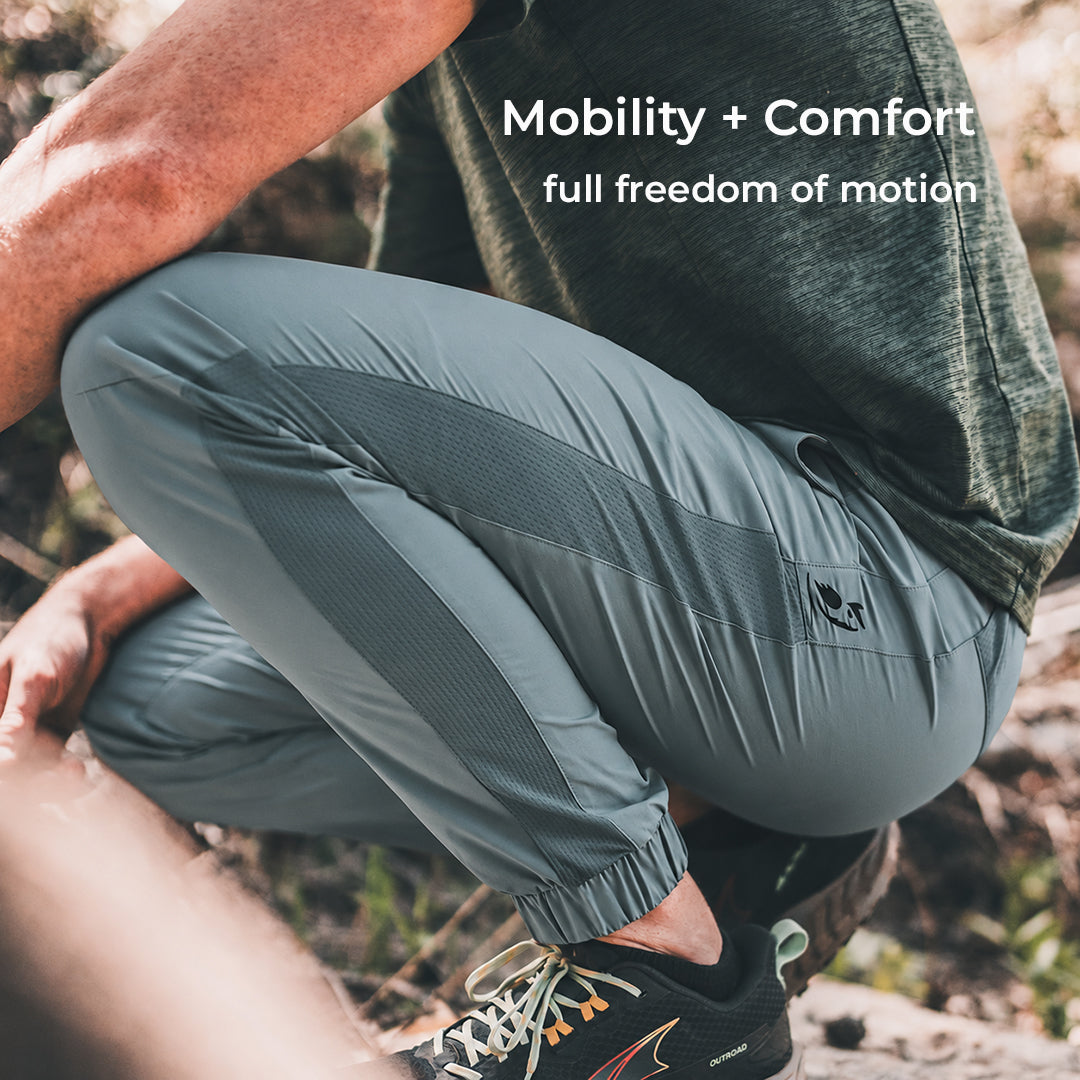 A person outdoors wearing blue athletic jogger-style pants with a small black logo near the lower leg, paired with a dark shirt and hiking shoes. White text at the top reads 'Mobility + Comfort full freedom of motion.