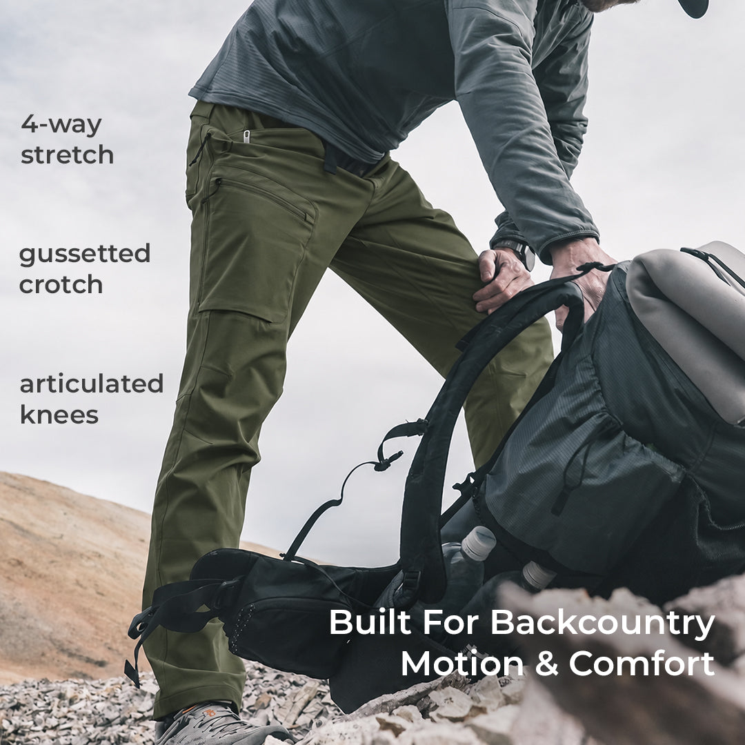 side view of man wearing green lightweight mountain pants on a peak while picking up a backpack
