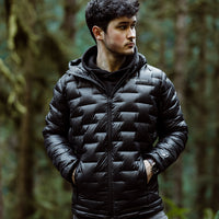 Person standing in a forest wearing a black Outdoor Vitals insulated jacket with hands in pockets, looking to the side.