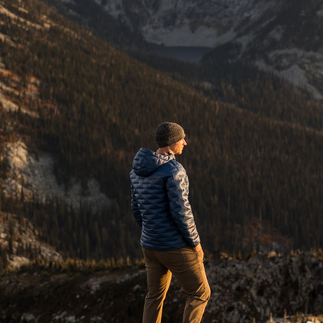 Person wearing a blue Outdoor Vitals insulated jacket and knit beanie, standing on a mountain ridge at sunset and looking out over a forested valley.