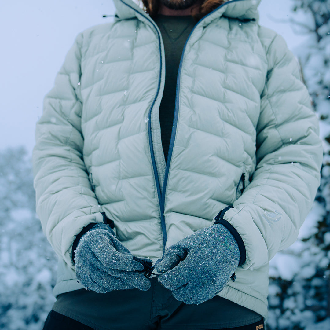 Person wearing a light gray Outdoor Vitals insulated jacket in snowy conditions, shown mid-zip with frosted gloves and visible snowflakes on the fabric.