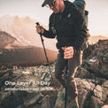 A man hiking on rocky mountains wearing a black one-layer jacket, brown pants, and a green backpack, with text overlay reading 'One Layer All Day comfortable from 25-50F'.